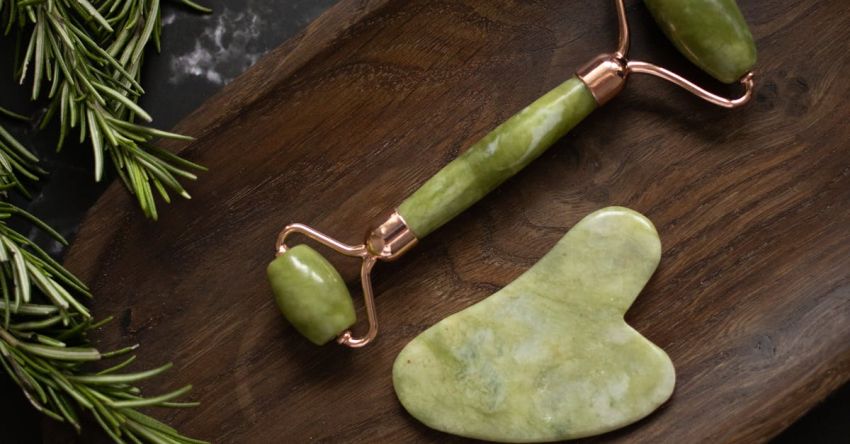 Muscle Definition - From above of Jade roller and Gua Sha massage tool placed on wooden plate on black marble table with fresh twigs of rosemary