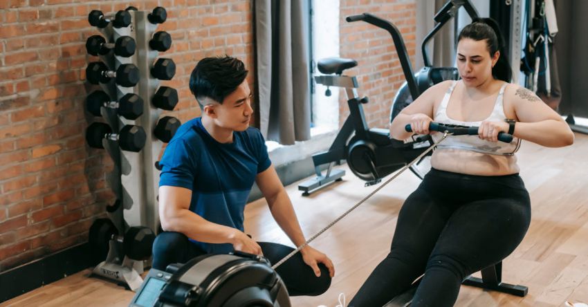 Rowing Machine - From above of professional ethnic constructor helping young plus size woman working out with rowing machine in sport club